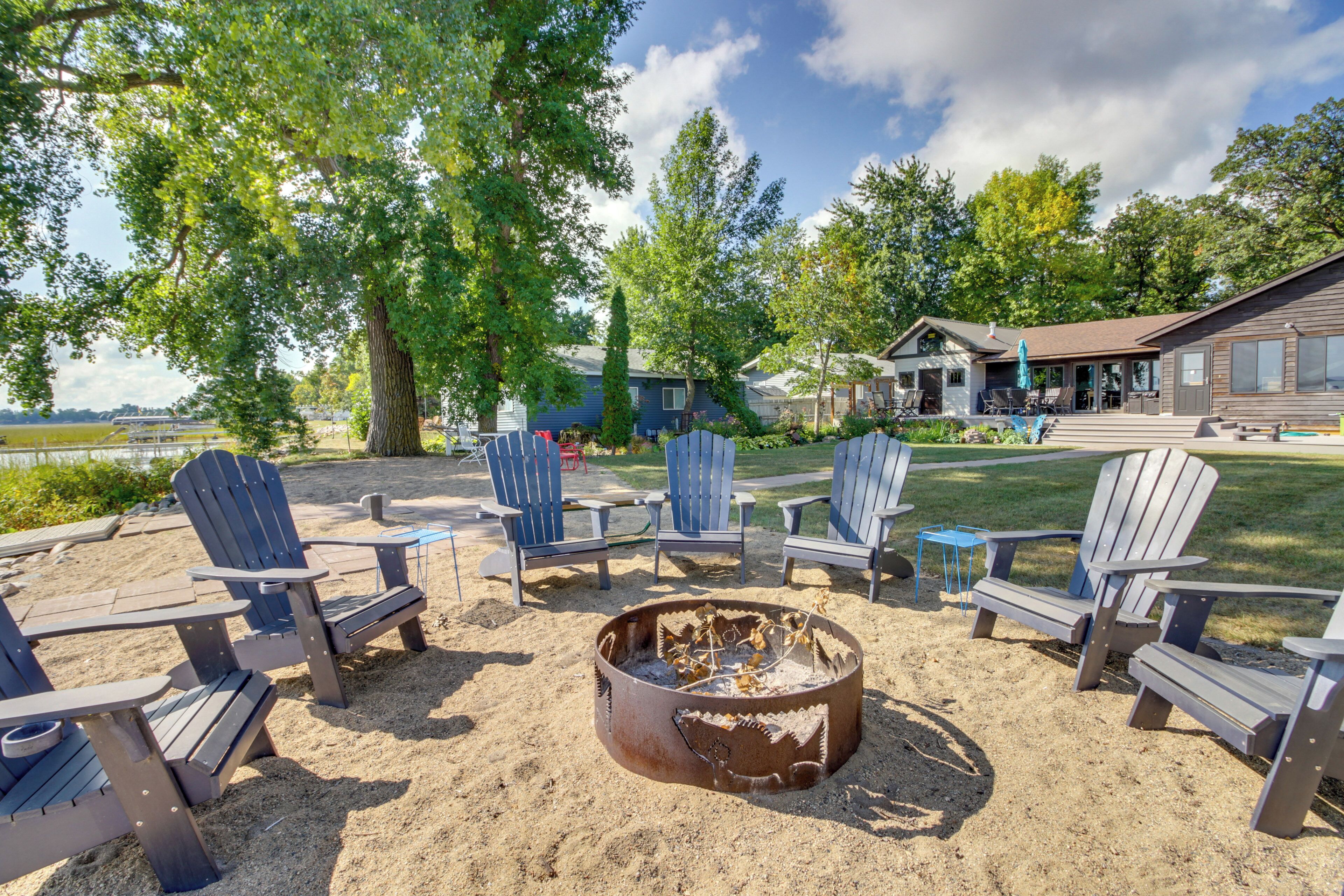 A beachside seating area with Adirondack chairs around a fire pit, a building with a porch, and a view of the water.