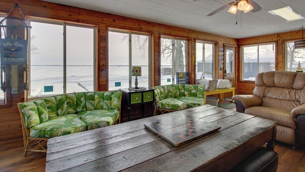 A cabin with a large window overlooking a lake, a ceiling fan, and a wooden table.