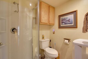 A bathroom with a glass shower enclosure, wooden cabinets, a framed picture on the wall, and a toilet with a paper towel holder. - Yellowstone Country Retreat: Stunning Views! (Livingston)