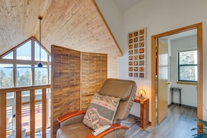 A cozy room with a wooden ceiling, a large window, a comfortable chair with a blanket, and a wall with framed pictures. - Angel Fire Retreat w/ Hot Tub ~ Hike In/out Skiing (Angel Fire)