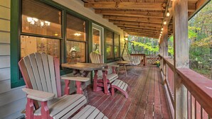 A covered porch with Adirondack chairs and a table, wooden beams, and large windows overlooking a forest. - Fletcher Cabin: Wraparound Deck & Pond Access (Fletcher)