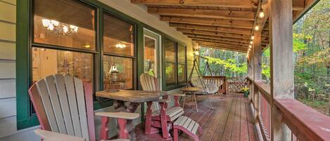 Eine überdachte Veranda mit Adirondack-Stühlen und einem Tisch, Holzbalken und große Fenster mit Blick auf einen Wald.