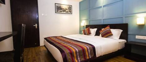 A hotel room with a wooden floor, a bed with a colorful striped blanket, a dark headboard, and a blue accent wall with a light fixture.
