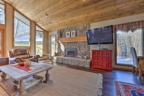 A cozy living room with a stone fireplace, a television mounted on the wall, and a large window offering a view of the outdoors. - Serene Ski Retreat - 3 Miles to Sun Valley Resort! (Sun Valley)