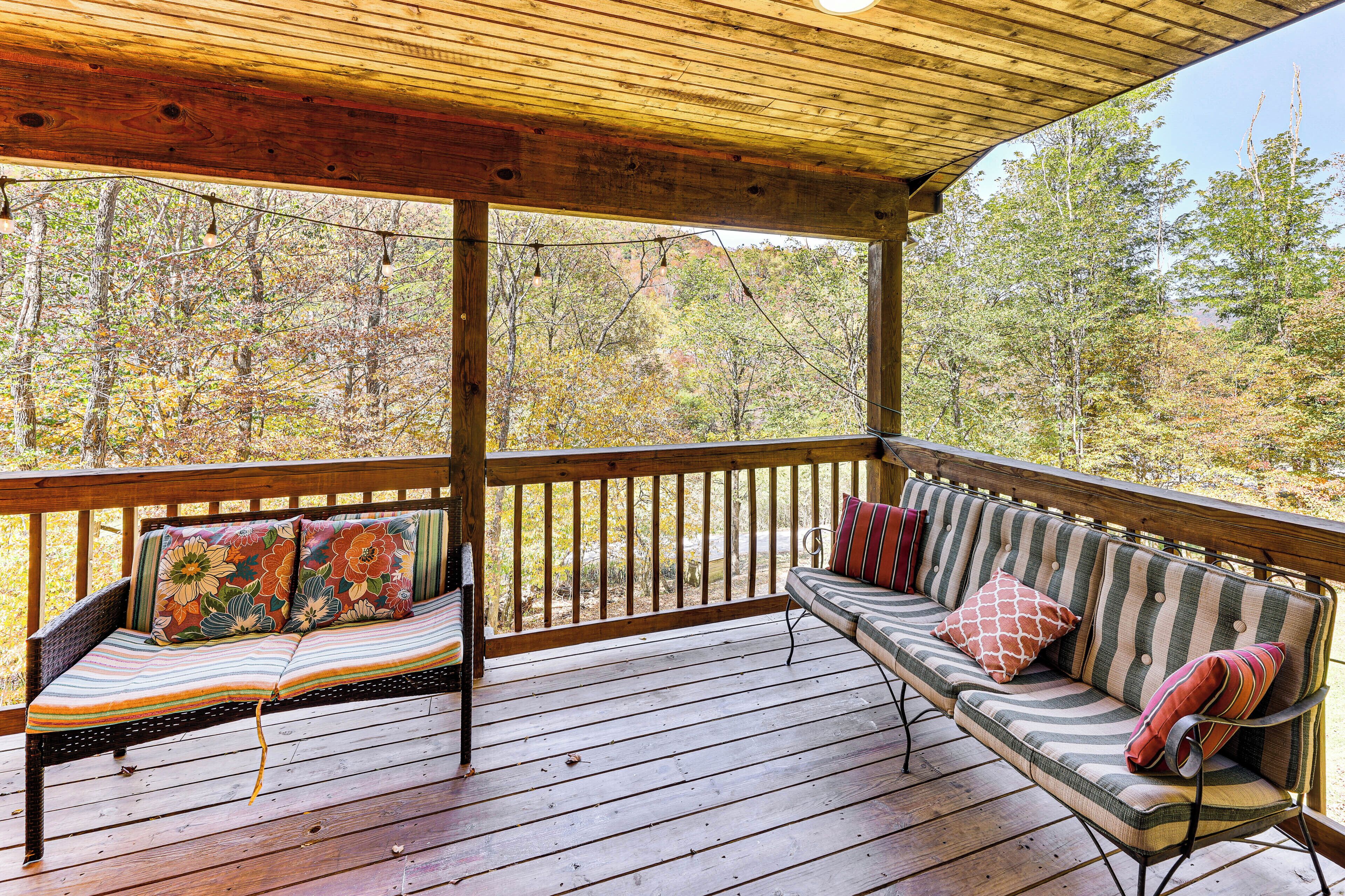 Eine Holzterrasse mit gepolsterten Sitzgelegenheiten, darunter eine Bank und eine Couch, beide mit gemusterten Kissen, mit Blick auf eine bewaldete Landschaft.