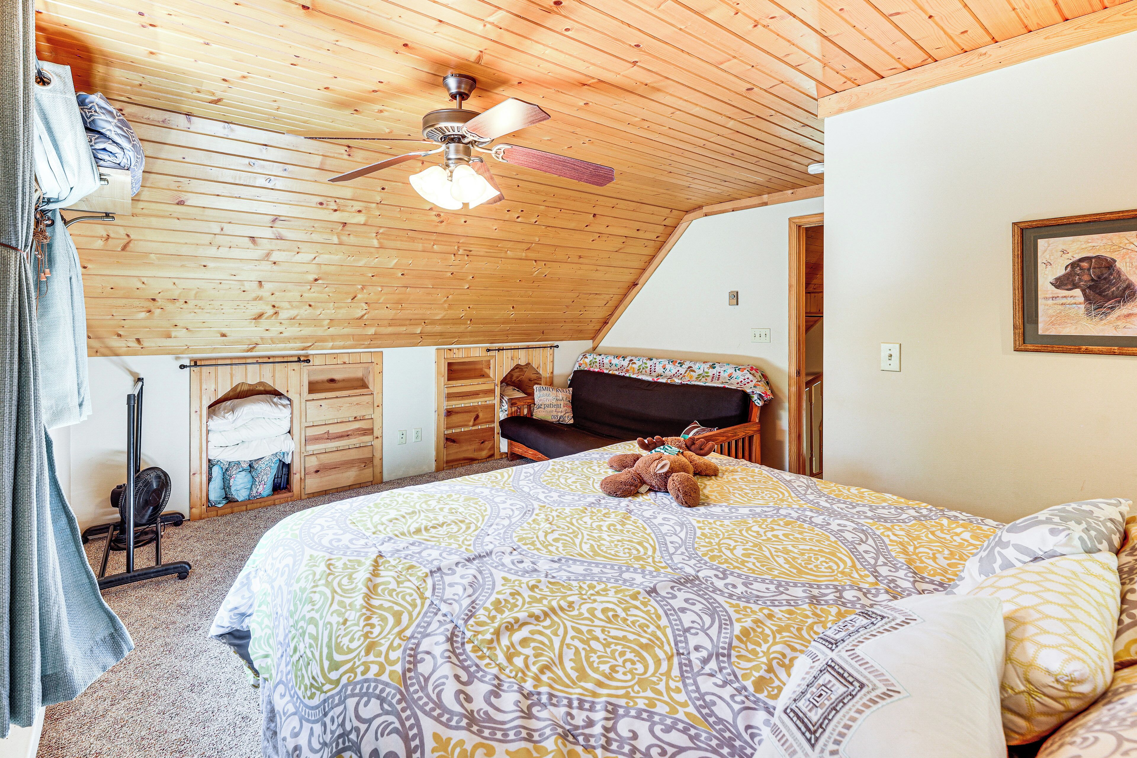Une chambre avec un plafond en bois, un lit recouvert d’une literie à motifs, un ventilateur de plafond et un tableau encadré accroché au mur.