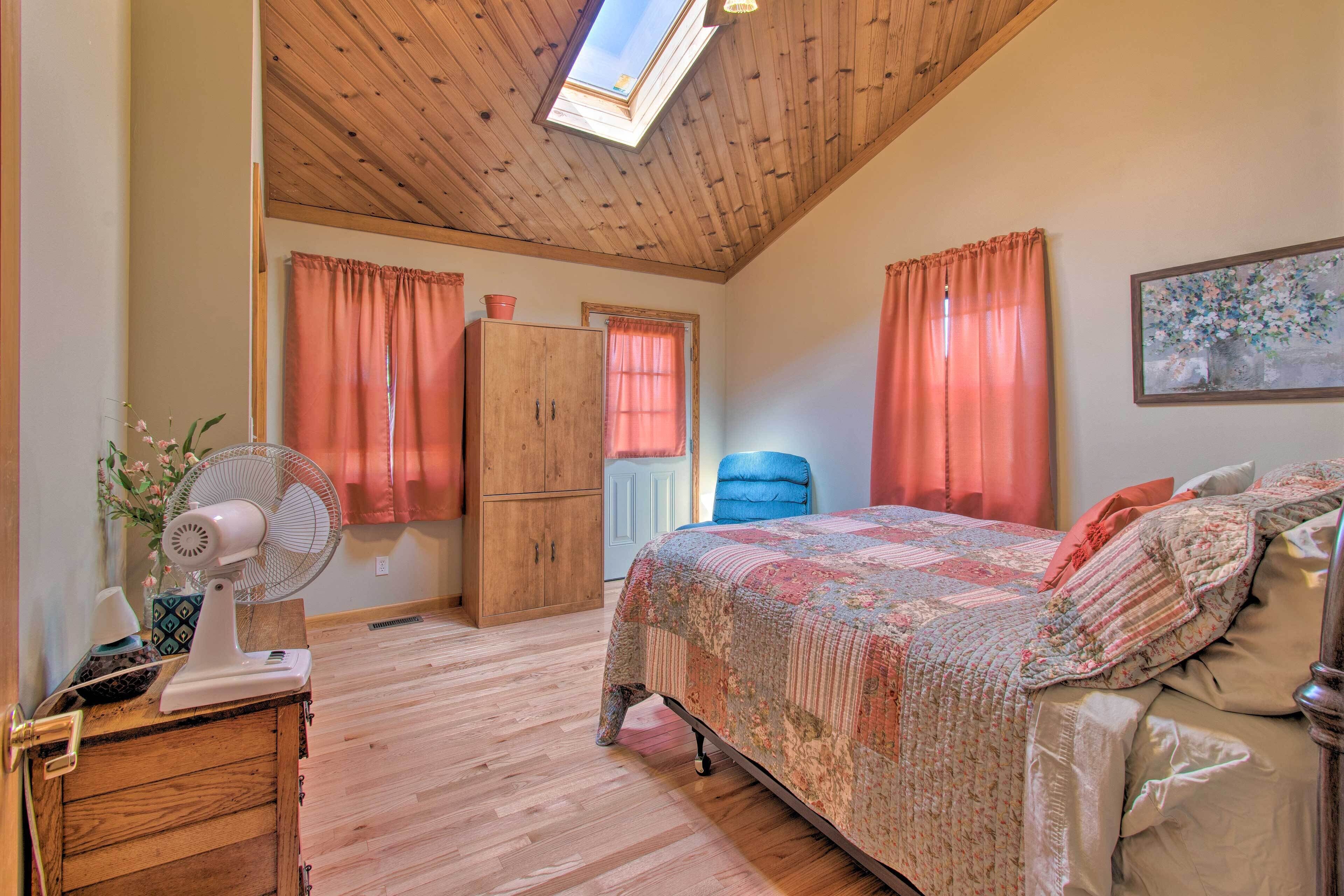 Een slaapkamer met een houten plafond, een bed met een bloemenprintdeken, een ventilator, een dressoir en een raam met gordijnen.