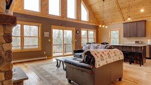 A spacious living room with a stone fireplace, large windows, a cozy sofa, and a kitchen area with wooden cabinets and a countertop. - Spacious Cabin: 10 Mi to Sunday River Skiing! (Bethel)