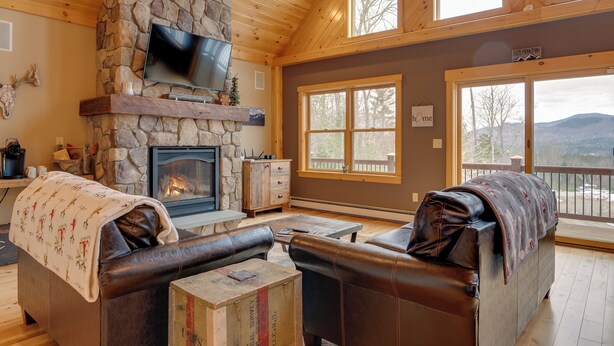 A cozy living room with a stone fireplace, leather sofas, and large windows offering a view of the mountains. - Spacious Cabin: 10 Mi to Sunday River Skiing! (Bethel)