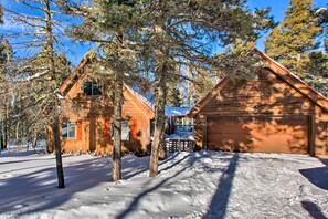 A wooden cabin with a garage, surrounded by snow-covered ground and trees. - Serene Angel Fire Cabin w/ Community Pools! (Angel Fire)
