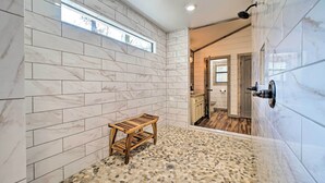 Un baño moderno con baldosas de mármol blanco, un banco de madera y suelo con diseño de guijarros.