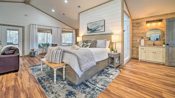 Une chambre à coucher avec un grand lit, une table de chevet, un banc, une fenêtre avec des rideaux, un plafond en bois et une salle de bain avec un lavabo et un miroir.