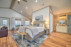 Un dormitorio con una cama grande, una mesita de noche, un banco, una ventana con cortinas, techo de madera y un baño con lavabo y espejo.