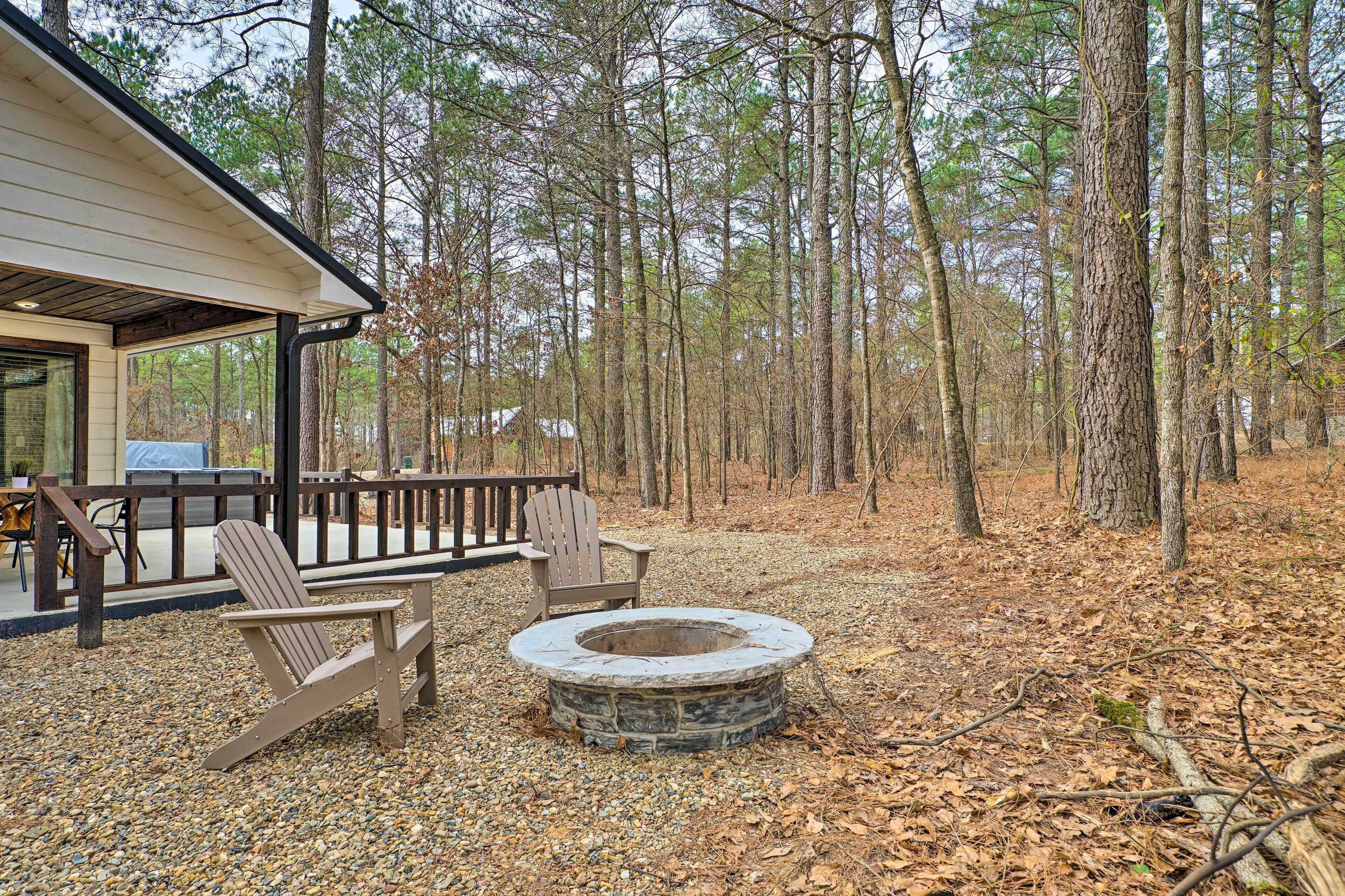 A cabin with a deck, outdoor seating, and a stone fire pit surrounded by a forest.