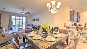 A dining area with a table set for six, surrounded by leather chairs, a chandelier, and a view of the living room and kitchen in the background.