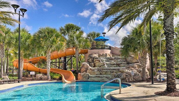 A swimming pool with a water slide, palm trees, and a rocky backdrop.