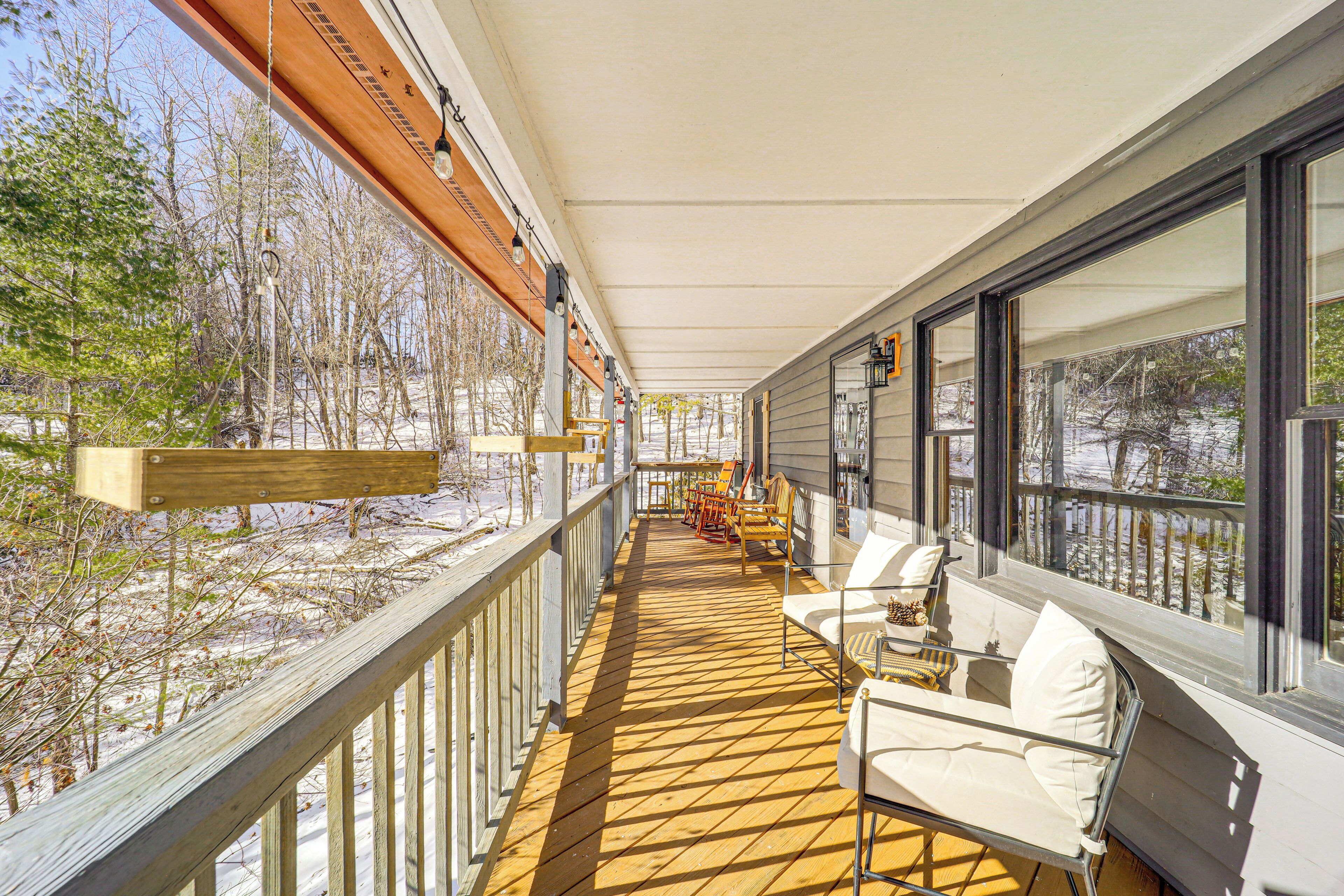 A covered outdoor deck with wooden railings, white outdoor furniture, and large windows offering views of a snowy landscape.