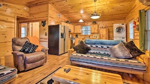 A cabin interior with a wooden ceiling, a sofa with patterned cushions, a recliner with a blanket, a wooden dining table, and a kitchen area with a washing machine. - Broken Bow Cabin w/ Hot Tub, Near Hiking + Fishing (Broken Bow)
