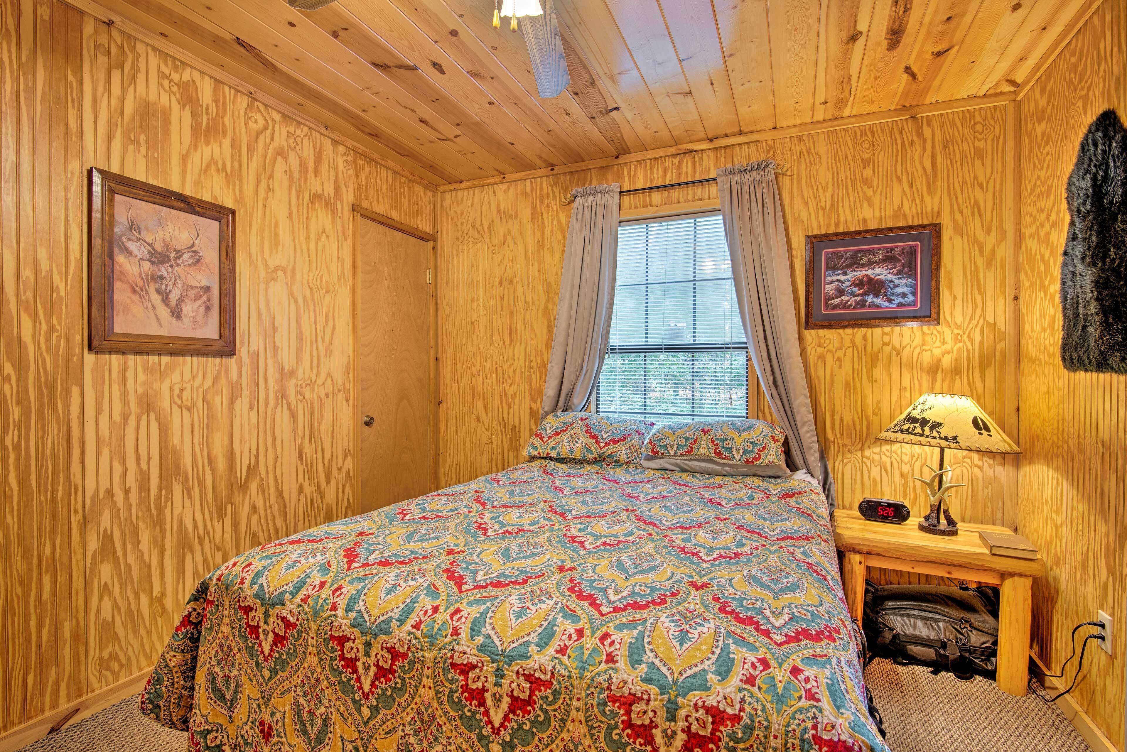 A wooden-paneled room with a bed, a window with curtains, a nightstand with a lamp, and two framed pictures.