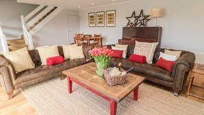 Ein Wohnzimmer mit braunem Ledersofa, einem Holztisch und einem roten Teppich. An den Wänden hängen gerahmte Bilder und eine Kristallleuchter ist angebracht.