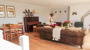 Ein Wohnzimmer mit einem braunen Sofa, einem Holzesstisch und Stühlen. Ein Klavier und ein Kamin mit Dekorationen an der Wand sind ebenfalls vorhanden.