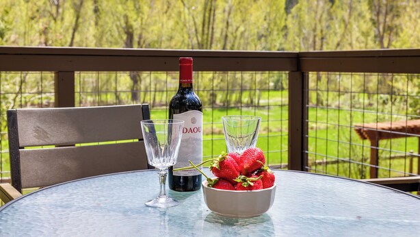 A table with a bottle of wine, two glasses, and a bowl of strawberries on a balcony with a view of a green landscape. - Furnished Deck & Nat'l Forest Views: Arnold Cabin (Arnold)