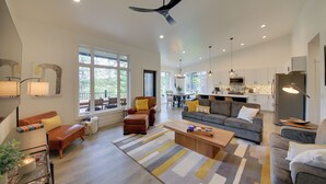 Amplia sala de estar con sofá, sillones, una mesa de centro y una mesa de comedor. Se observa una cocina al fondo, equipada con electrodomésticos modernos y un ventanal grande que proporciona luz natural.