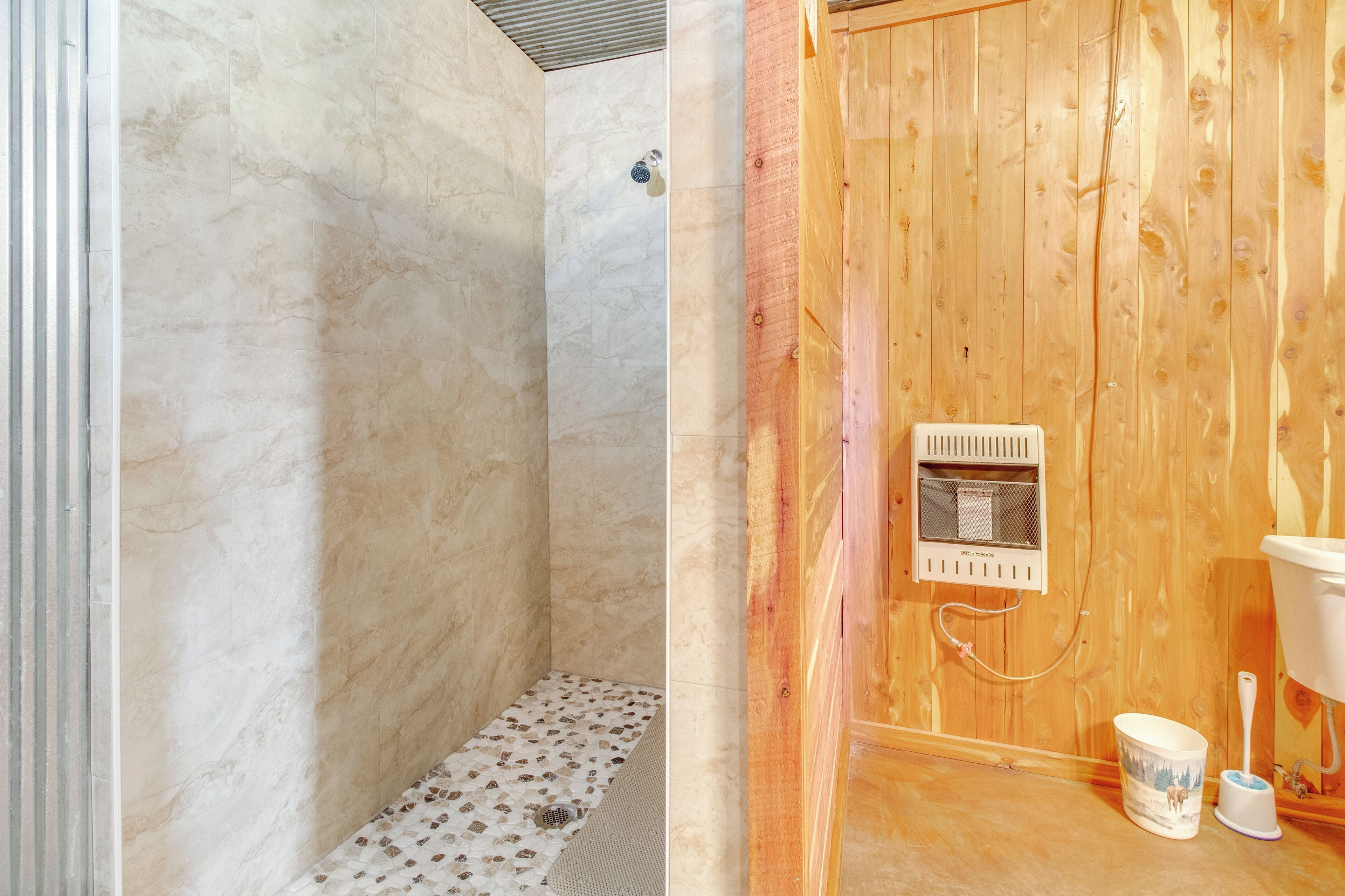 A bathroom with a wooden wall, a shower with a marble floor, and a toilet with a bucket and brush.