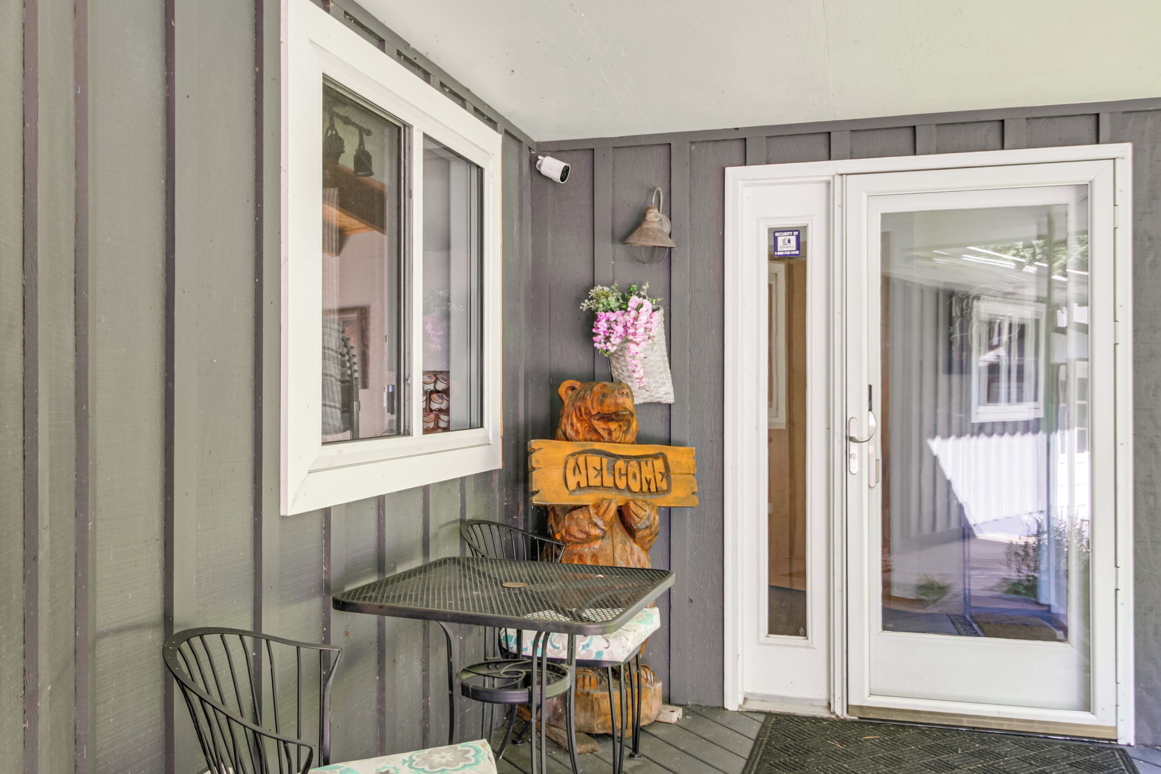 Eine Veranda mit Tisch und Stühlen, ein Holzschild mit der Aufschrift 'WELCOME' und eine Tür mit Glasfenster.