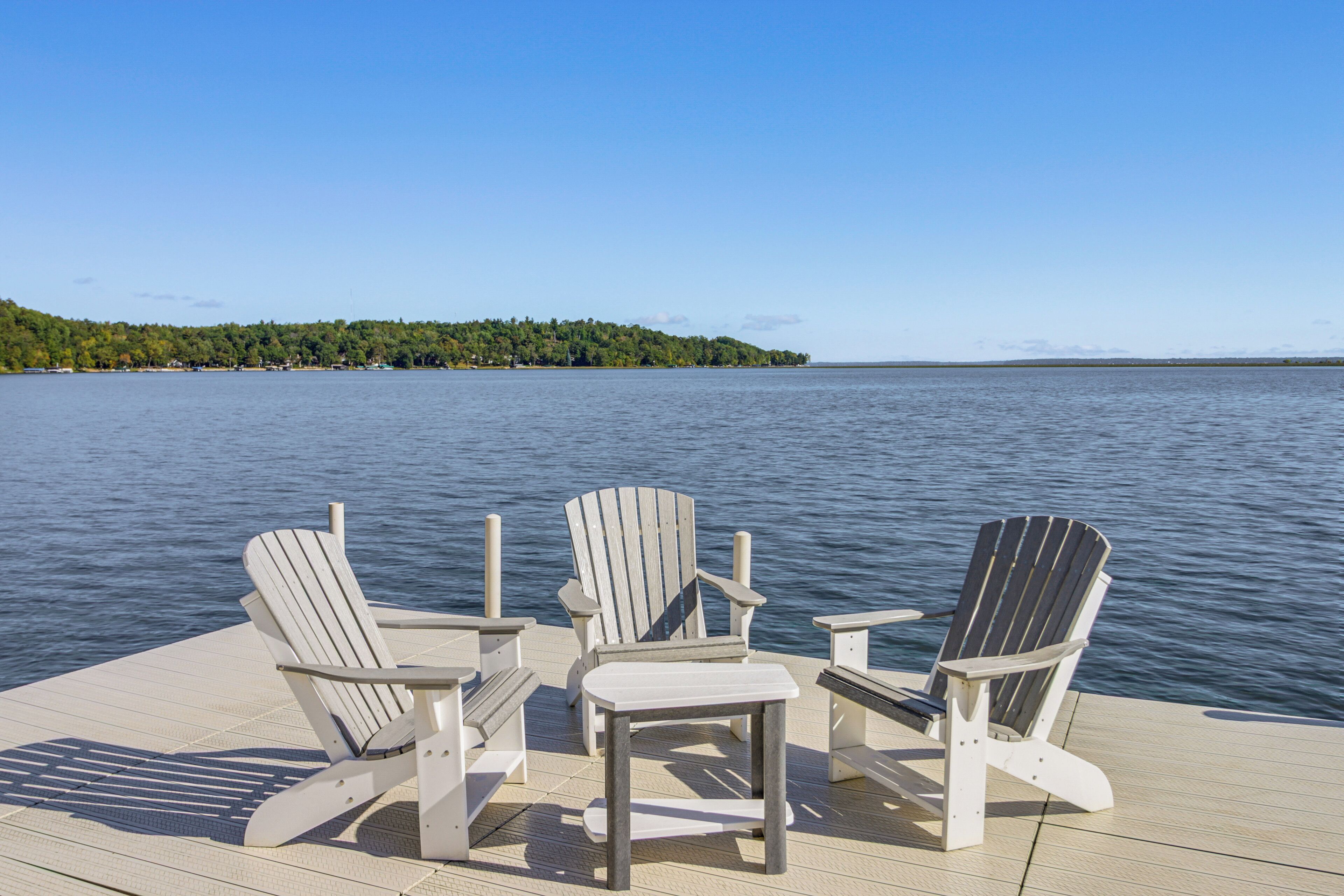 Three Adirondack chairs on a wooden deck with a view of a calm lake and a forested island in the distance.