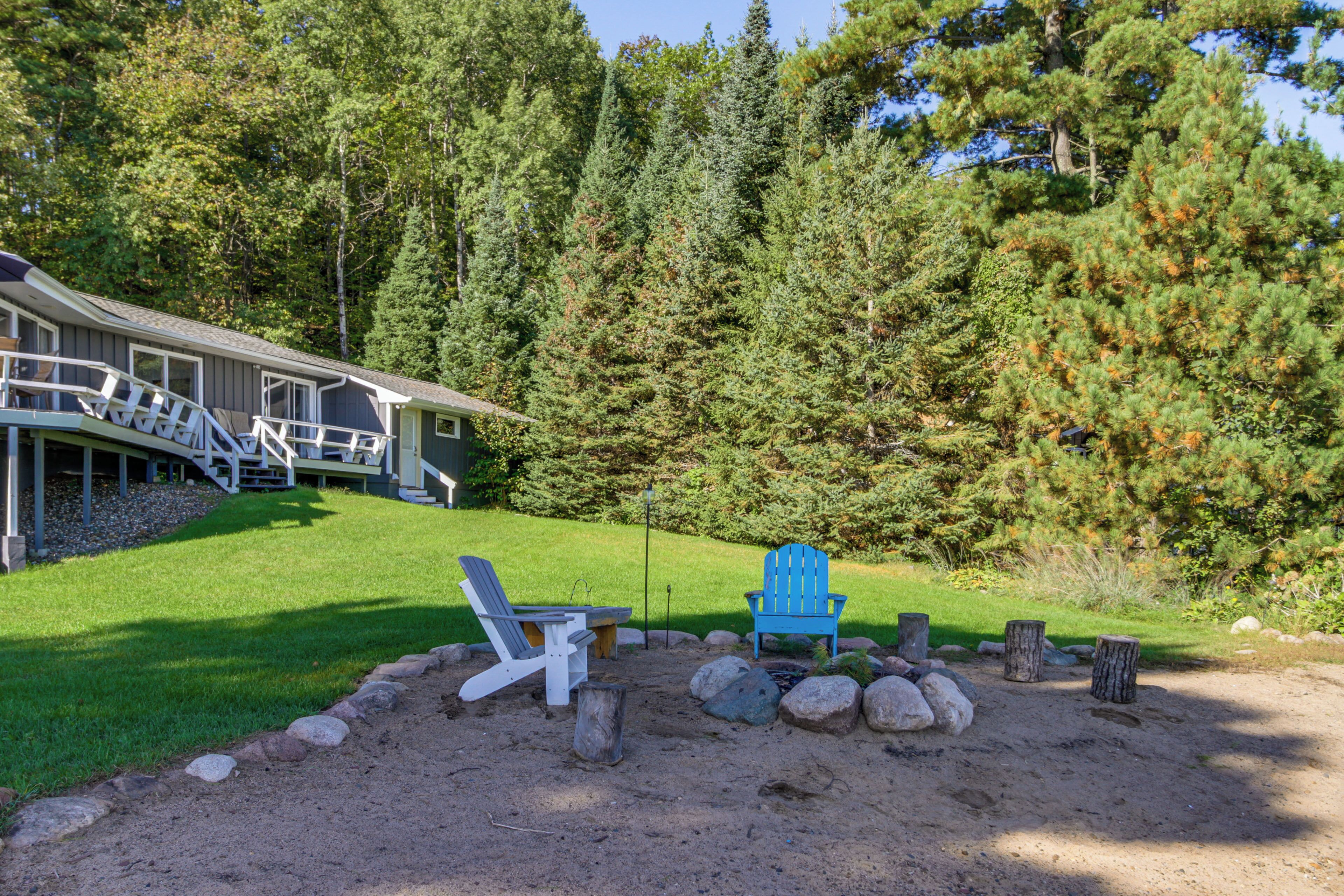 A house with a deck, two Adirondack chairs, and a fire pit in a forested area.