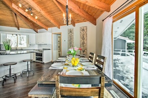 A dining area with a wooden table set for four, a kitchen in the background, and a large window with a view of a snowy landscape. - Waterfront Gull Lake w/ Dock: Family Friendly! (Nisswa)