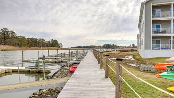 A wooden pier leading to a dock with boats, a grassy area with colorful kayaks, and a building with balconies.