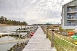 A wooden pier leading to a dock with boats, a grassy area with colorful kayaks, and a building with balconies. - Bright Lewes Condo w/ Balcony, 7 Mi to Beach! (Lewes)