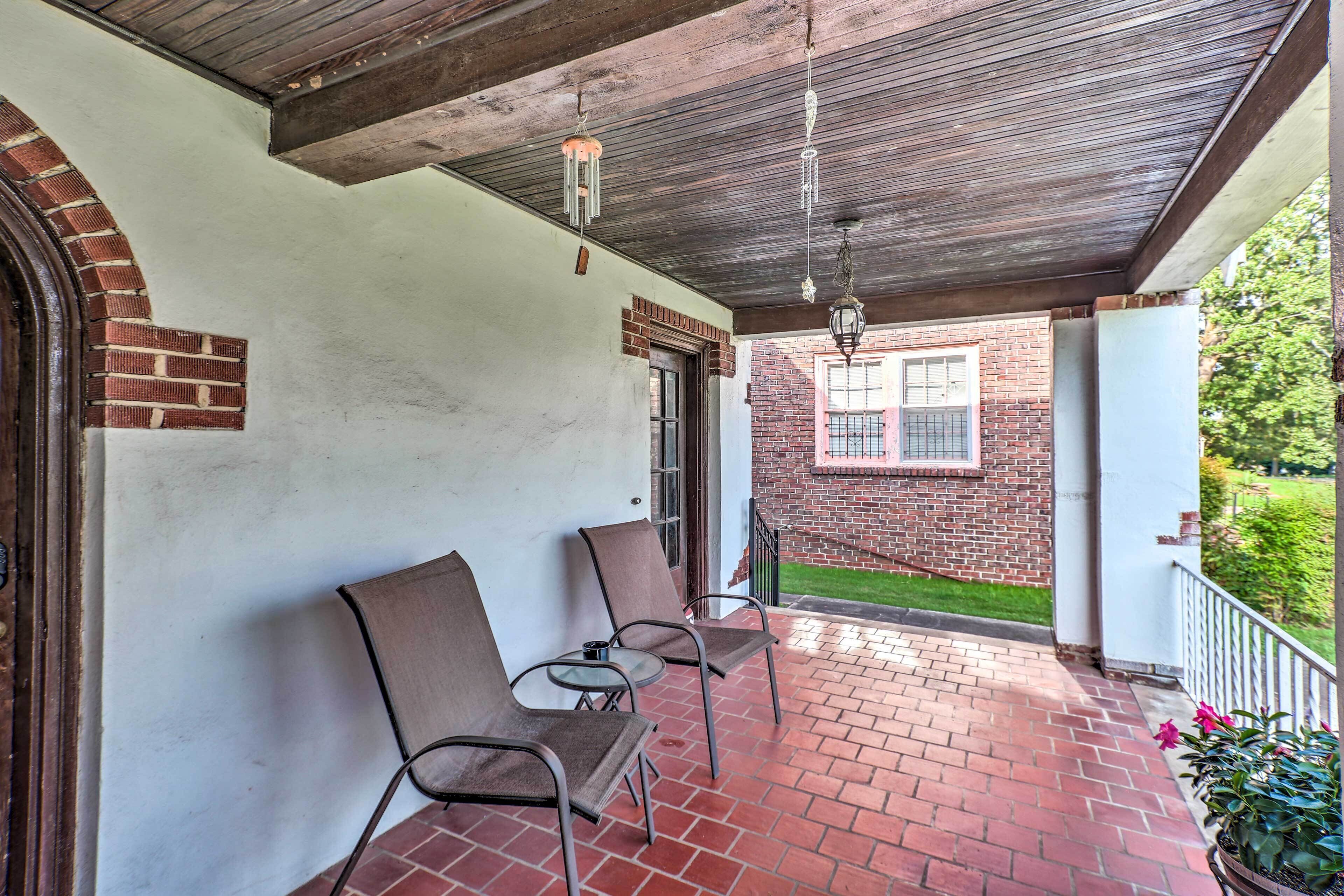 Un porche abrité avec deux chaises et une table, un mur en briques et une fenêtre donnant sur un jardin.