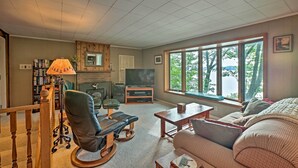 A living room with a fireplace, a television, a sofa, and a large window with a view of trees. - 2 Mi to Winery: Le Roy Escape w/ Fire Pit & Views (Le Roy)