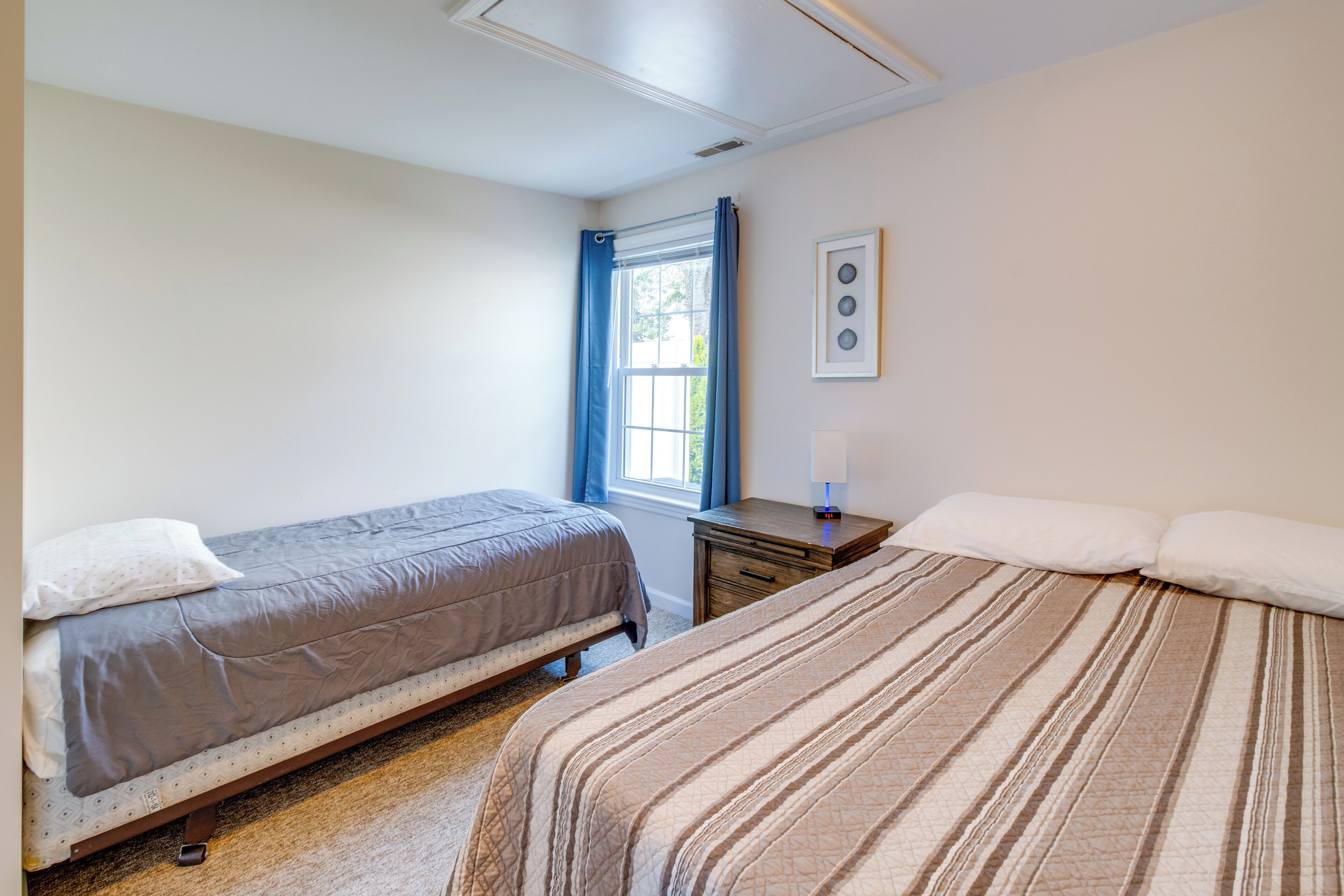 A bedroom with two beds, a window with curtains, a bedside table with a lamp, and a framed picture on the wall.