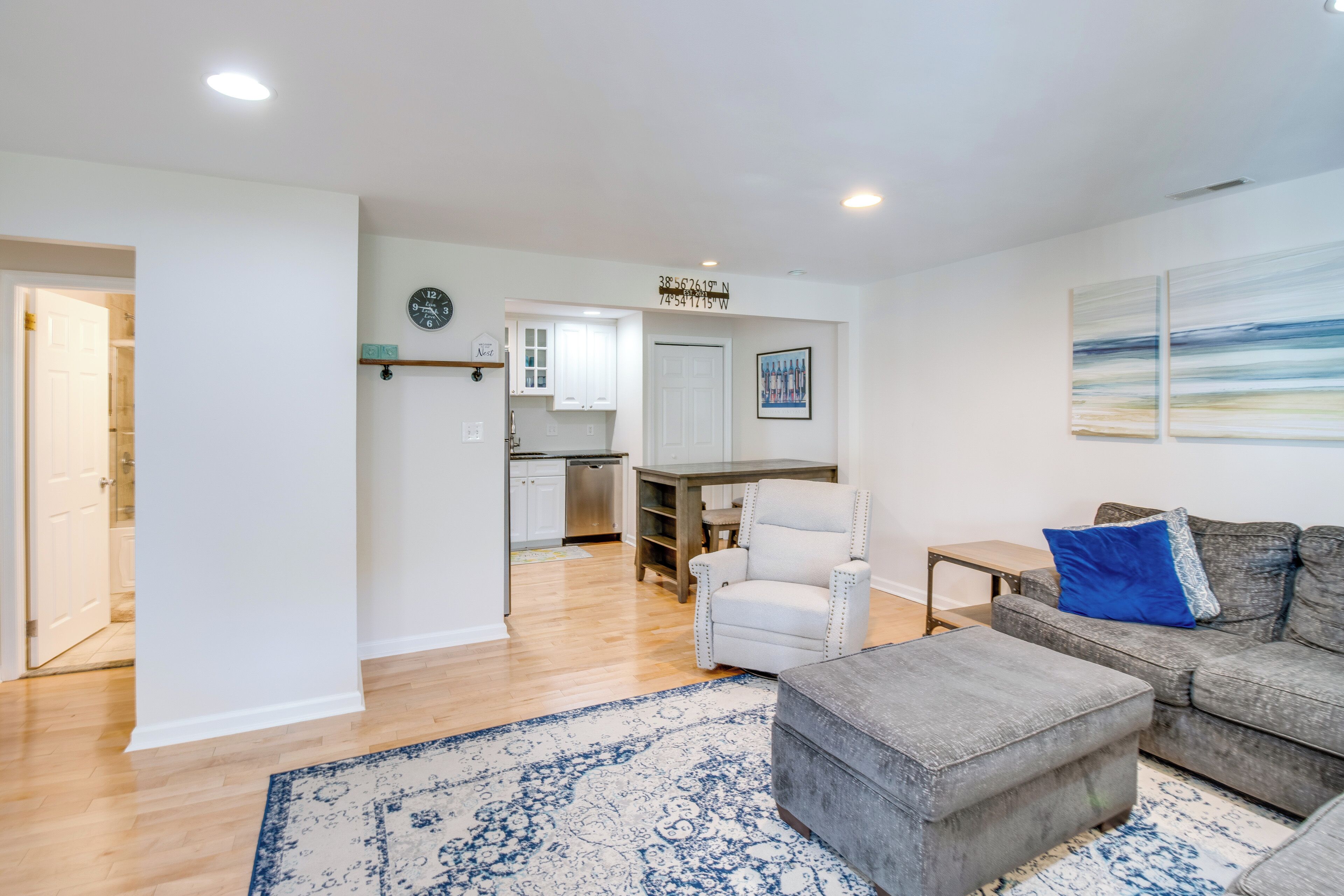 A spacious living room with a grey sofa, a white armchair, and a blue patterned rug. A kitchen with white cabinets and a dining area with a table and chairs are visible in the background.