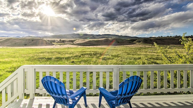 A deck with white railings and two blue chairs facing a grassy field with mountains in the background.