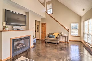 A spacious living room with a fireplace, a flat-screen TV mounted above it, a cozy sofa, and a staircase leading to an upper level. - Heber Springs Cabin ~ 1 Mi to Sugarloaf Mtn! (Heber Springs)