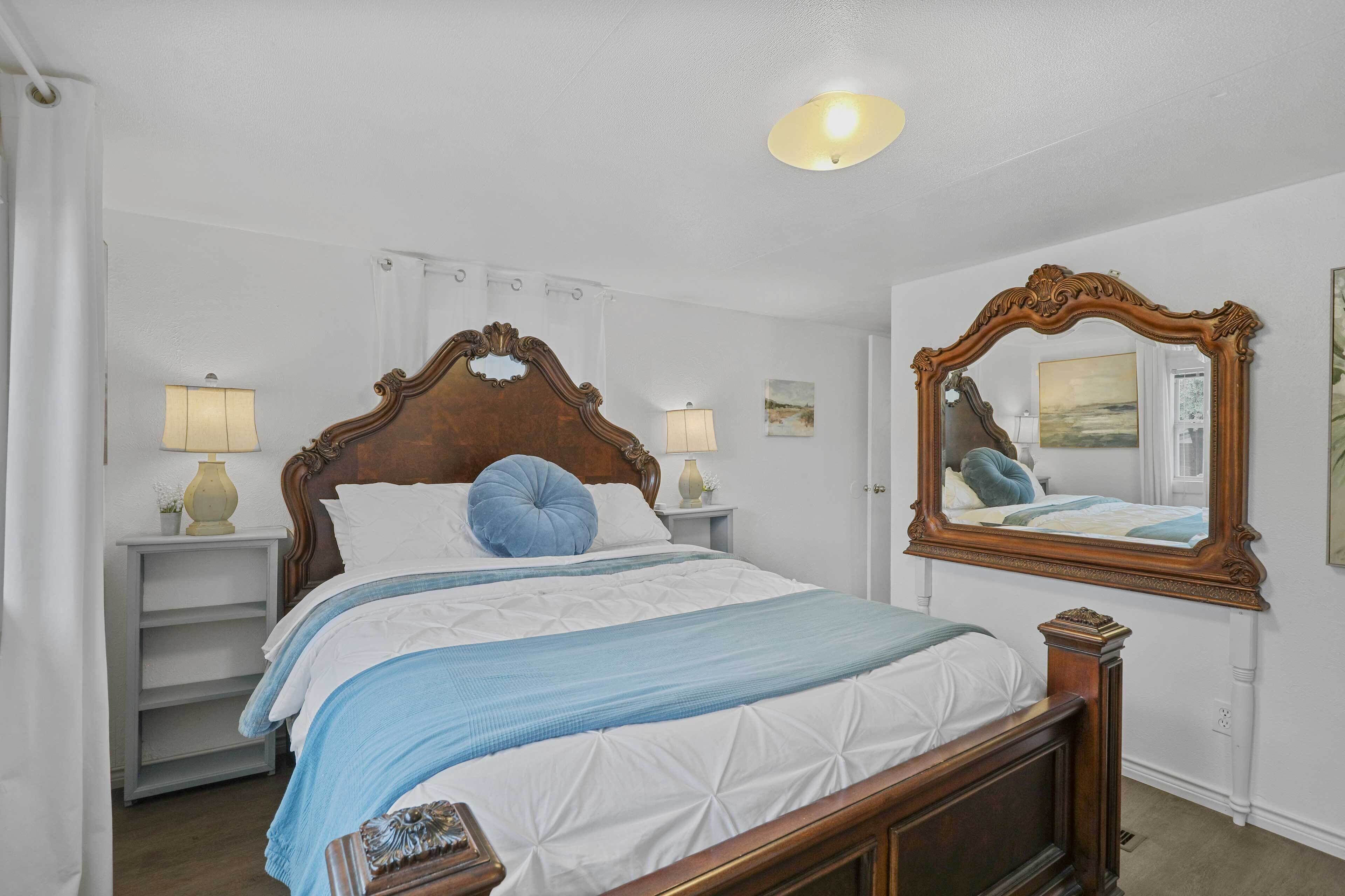 Une chambre à coucher bien rangée, avec un lit en bois, deux tables de chevet avec des lampes, un grand miroir et un tableau encadré accroché au mur.