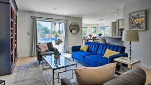Una sala de estar moderna con un sofá azul, una mesa de centro de vidrio y un sofá gris. Hay una estantería con luces anaranjadas, un área de comedor al fondo y un ventanal con vistas a árboles.