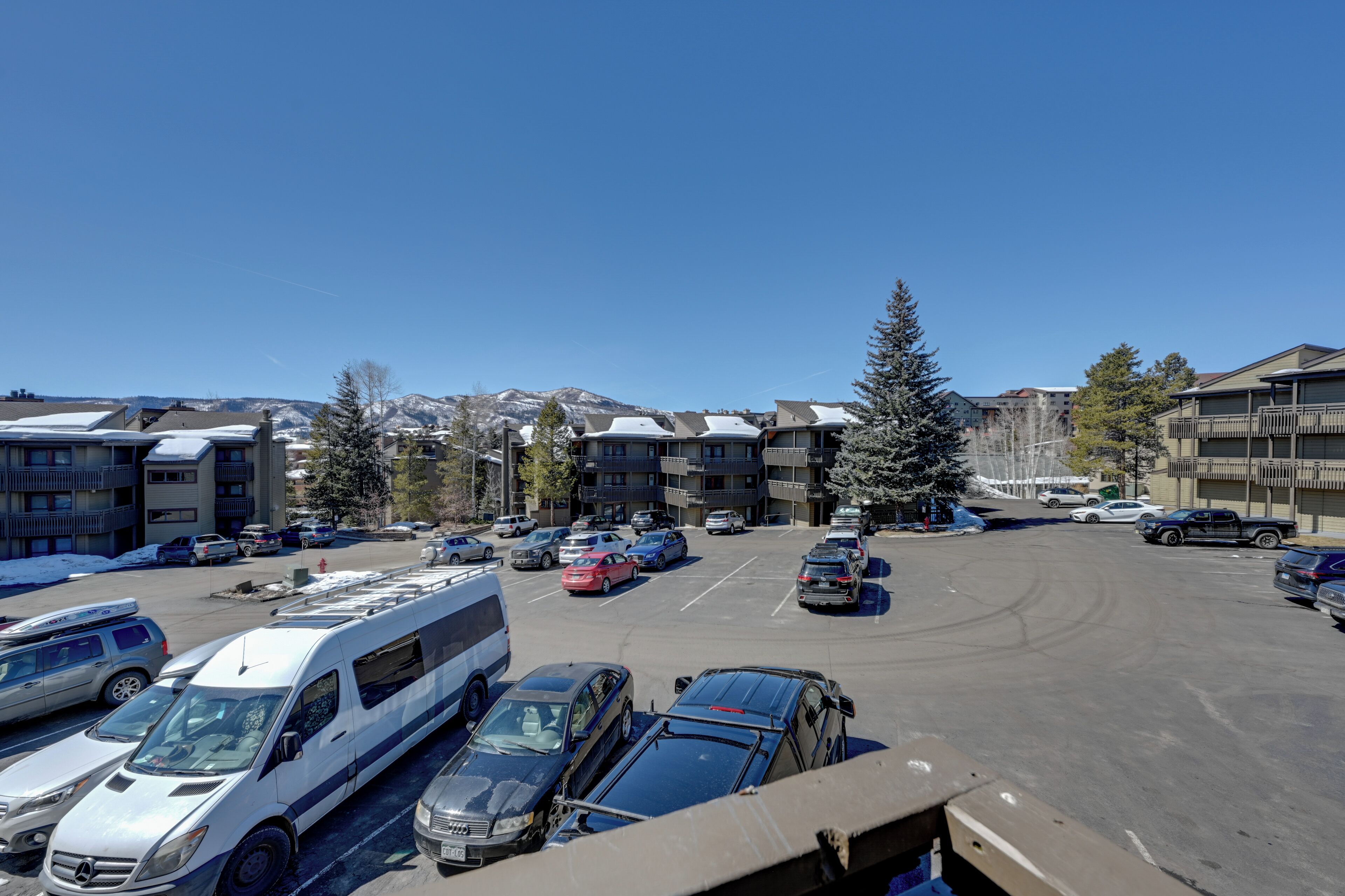 Un parking où sont garées plusieurs voitures et un autobus, entouré d’immeubles dotés de balcons et d’un grand arbre à feuilles persistantes.