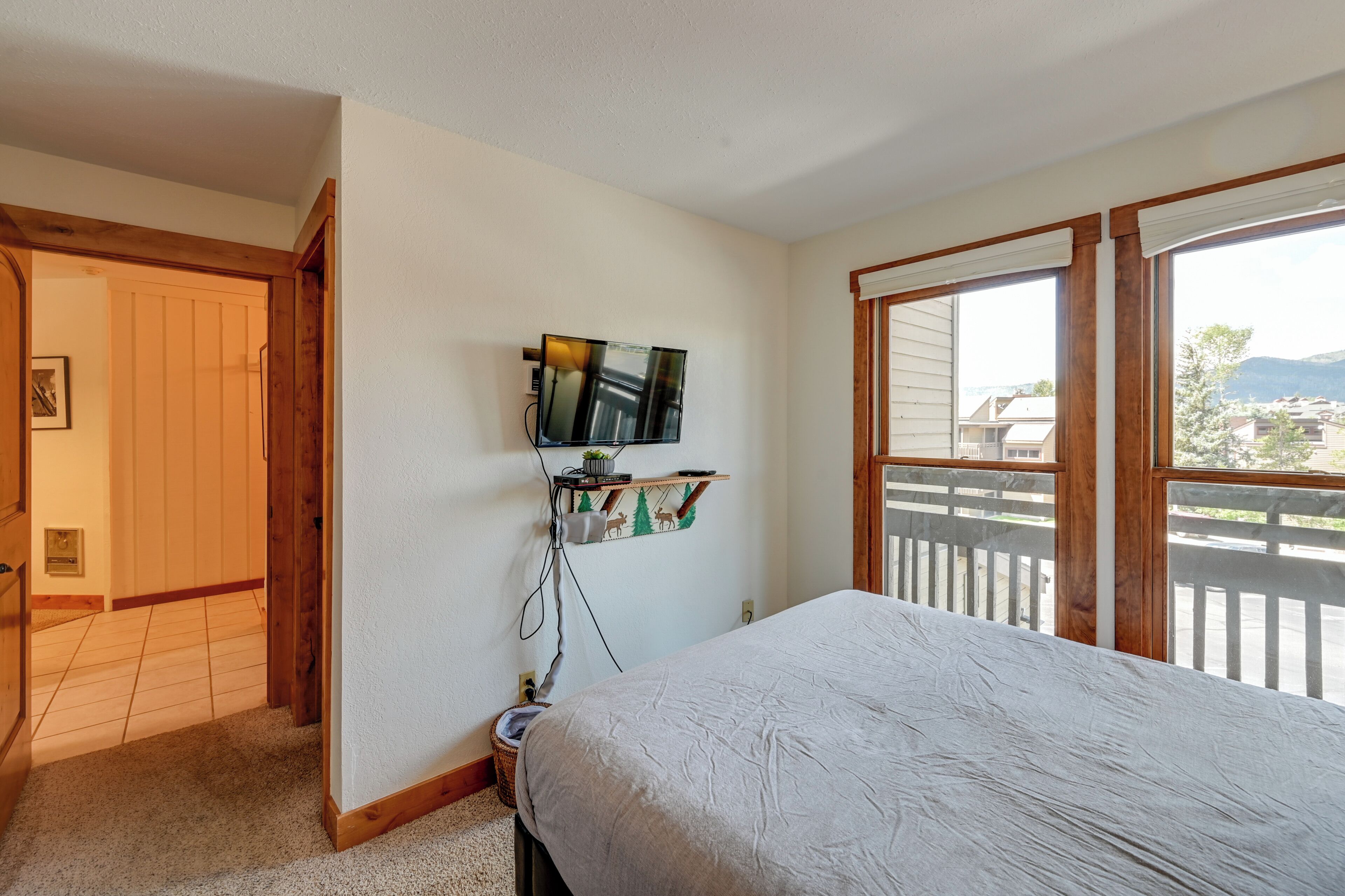 Une chambre à coucher comprenant un lit, un téléviseur fixé au mur, une petite étagère avec des objets décoratifs et un balcon offrant une vue sur les bâtiments et les montagnes.