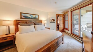 Een slaapkamer met een houten bedframe, een nachtkastje met een lamp, een schilderij aan de muur, en een groot raam met uitzicht op het omliggende landschap.