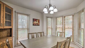 Un coin repas avec une grande table, des chaises en bois et un lustre. De larges stores blancs habillent les fenêtres, et un tableau est visible au mur.