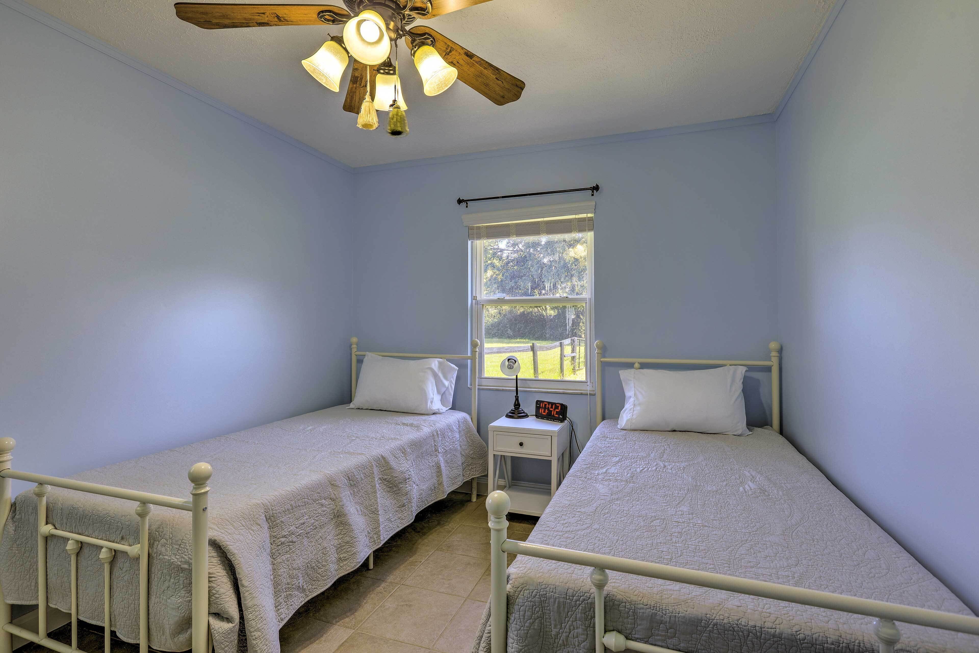 A bedroom with two single beds, a ceiling fan, a window with a view of greenery, and a bedside table with a clock.