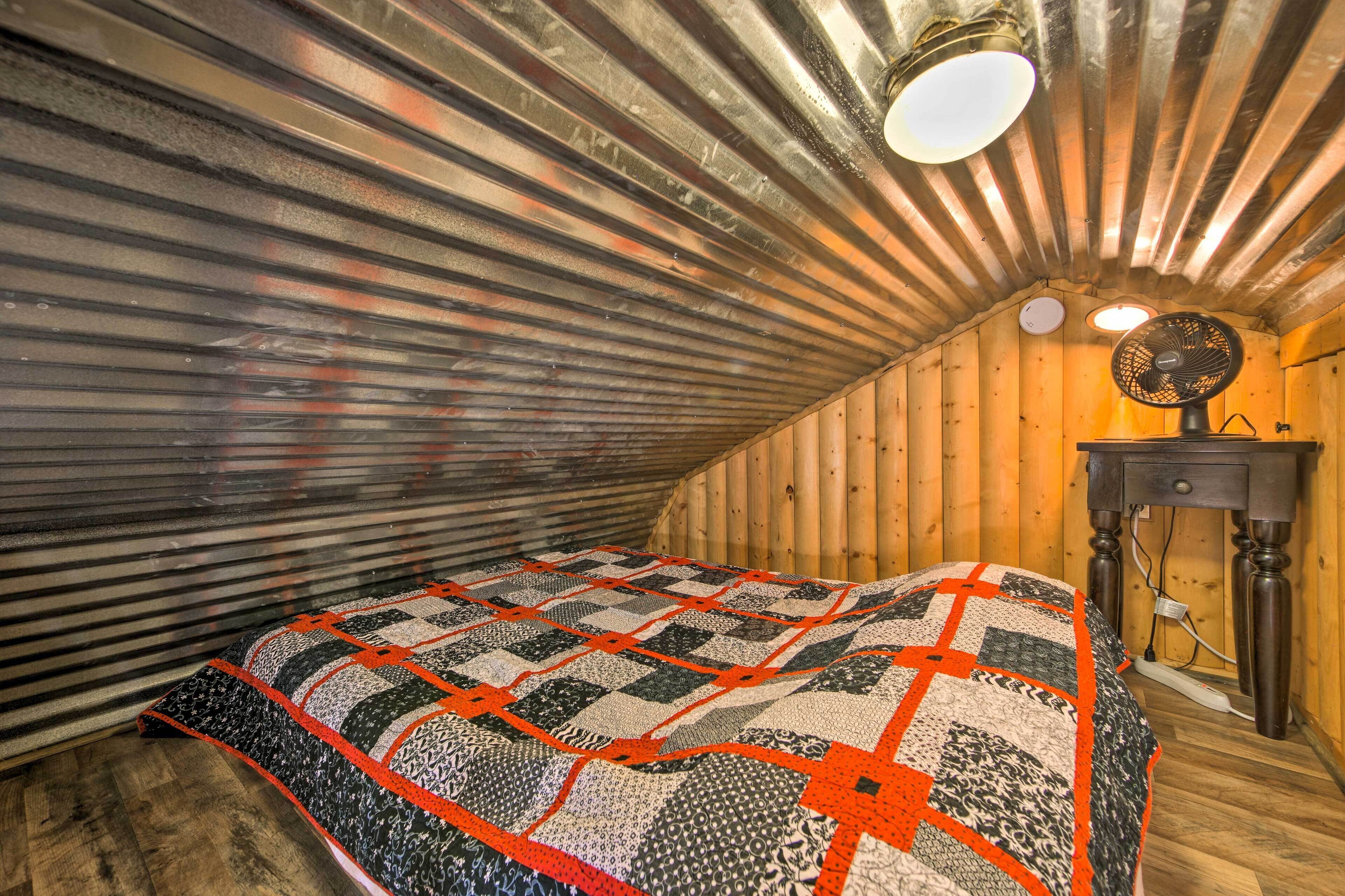 Un dormitorio acogedor en el ático, con techo inclinado, colcha acolchada, cómoda de madera y ventilador de pared.