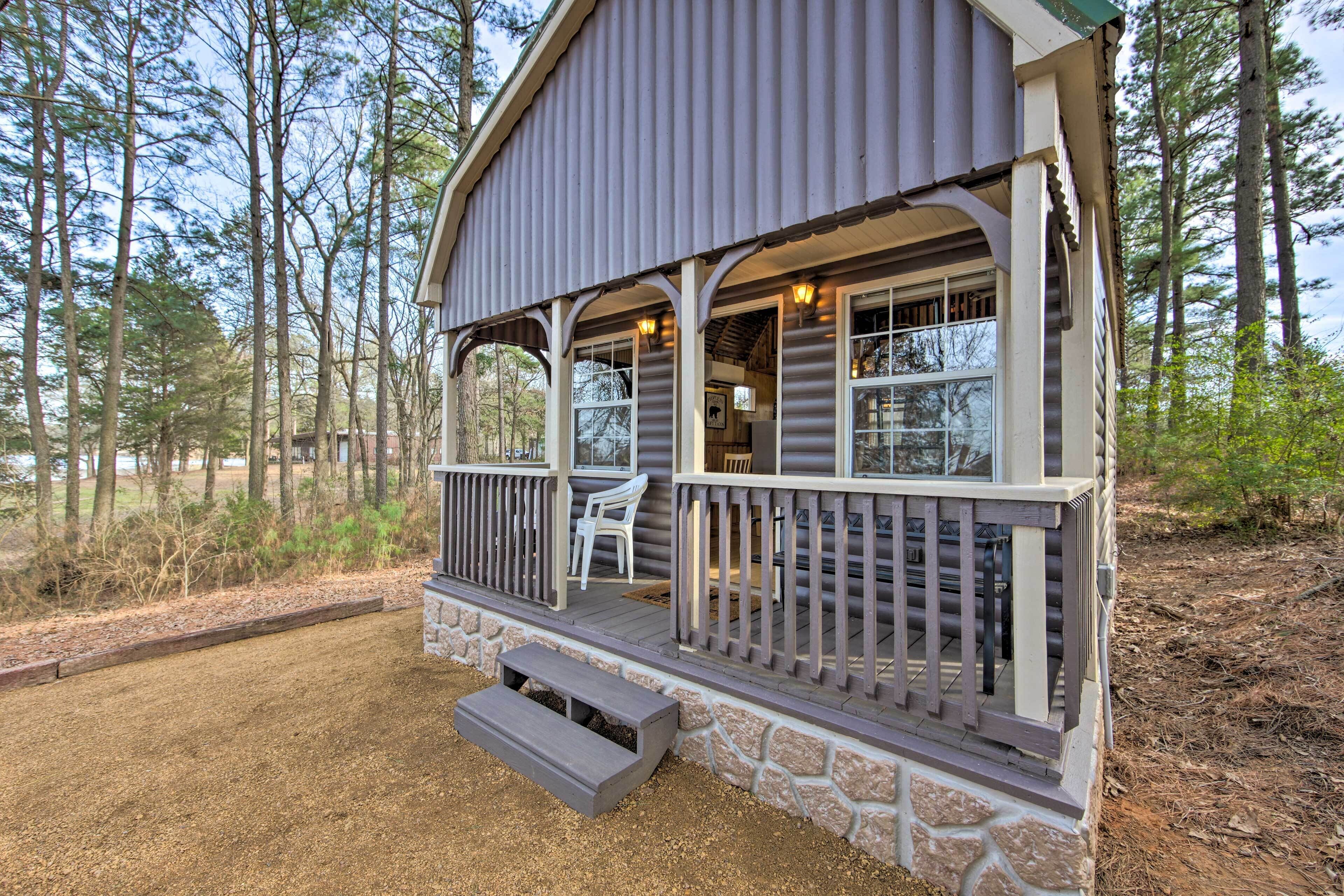 A log cabin with a porch, stone foundation, and a view of the surrounding forest.