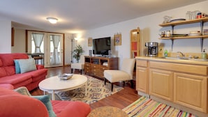 Una sala de estar con un sofá rojo, una mesa de centro blanca y un televisor. Hay una zona de cocina con armarios de madera y un lavabo.